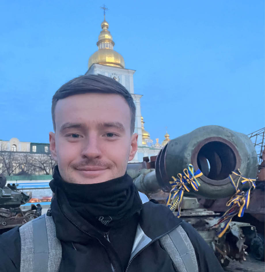 A young man wearing a scarf, standing outside, smiling, with a blue sky behind him