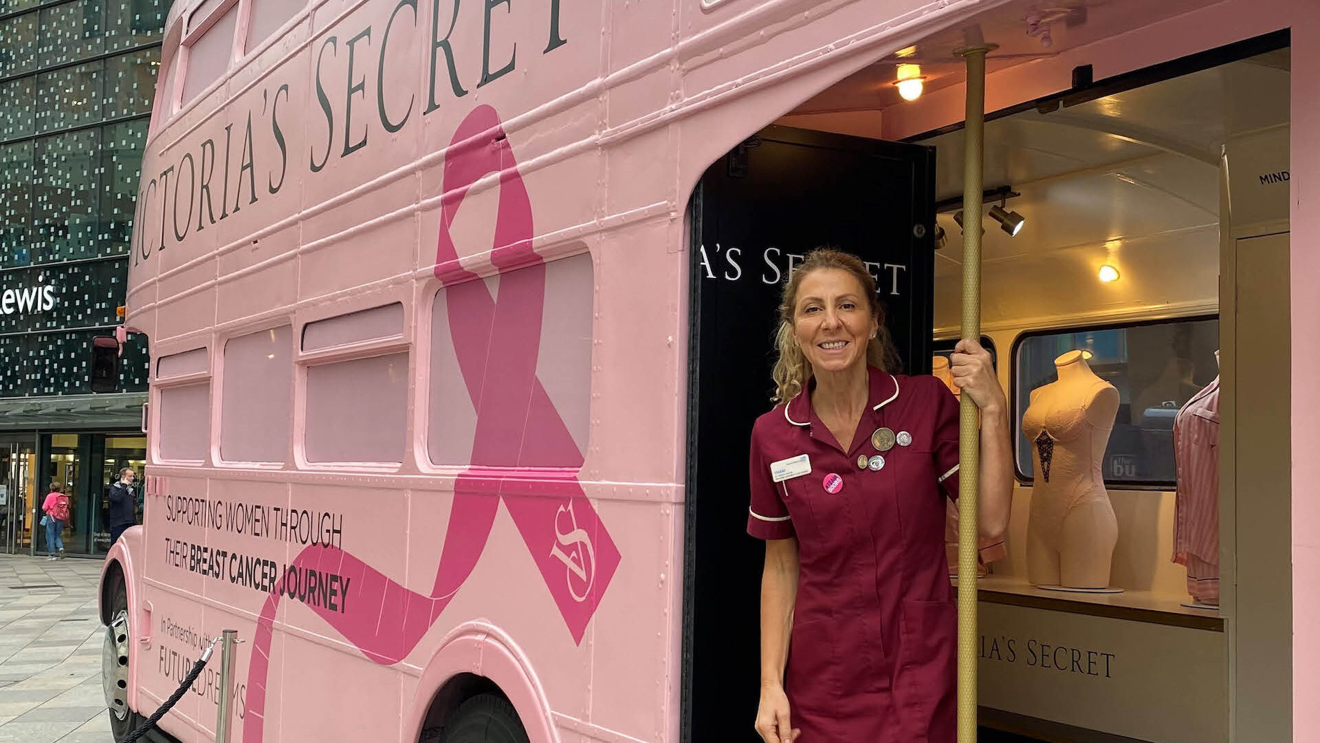 Outside shot of a woman in a nurse uniform standing on the bottom deck of a pink Breast Cancer bus